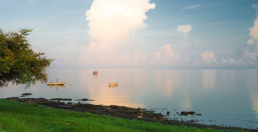 Mar en calma al amanecer en el malecón de Campeche, como un espejo que refleja las nubes; dos pequeñas barcas descansan cerca de una orilla de piedras y pasto verde, junto a un árbol.