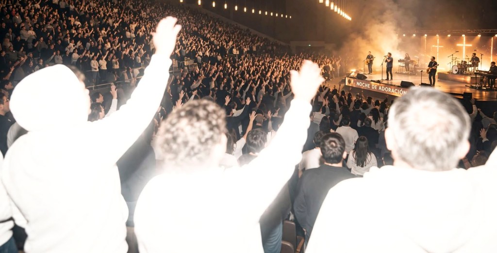 Recreación de una foto con flash en un auditorio durante una noche de adoración: las personas en primer plano, con las manos levantadas, aparecen blancas y brillantes por la sobreexposición, mientras el escenario al fondo se ve con su iluminación normal.